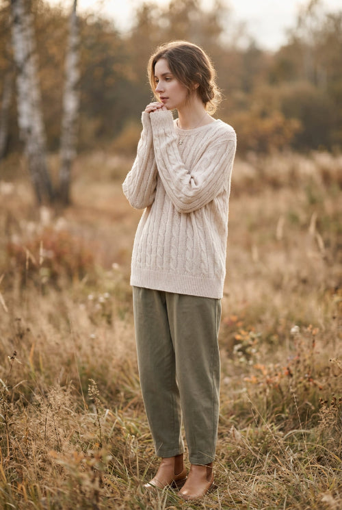 A person wearing a cream-colored cable-knit sweater with a crew neck and ribbed hem, paired with beige pants, photographed indoors against a light wall.