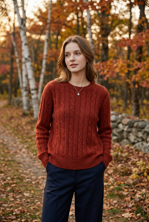 A person wearing an oversized cable-knit sweater in a warm rust red color, paired with dark pants, holding a phone in front of their face.
