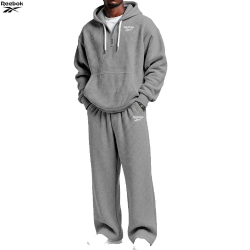 A man wearing a gray waffle-knit hoodie and matching sweatpants set from Reebok, shown against a plain white background. The Reebok logo is visible on the chest and thigh of the clothing.