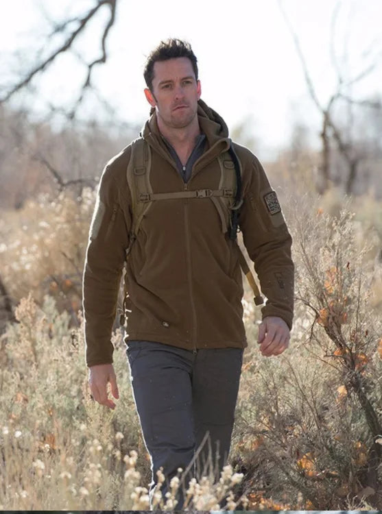 Man walking outdoors in a natural setting wearing a brown jacket and carrying a backpack.
