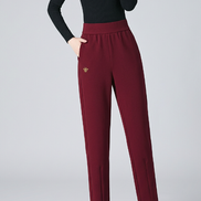 A woman modeling high-waisted, straight-leg pants in a deep wine red color, paired with a black long-sleeve top and black heels. The image features a dark gray studio background .