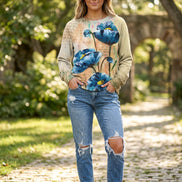 A woman wearing a beige crewneck sweatshirt with a large blue poppy floral print, paired with blue jeans, holding a bouquet of white roses.