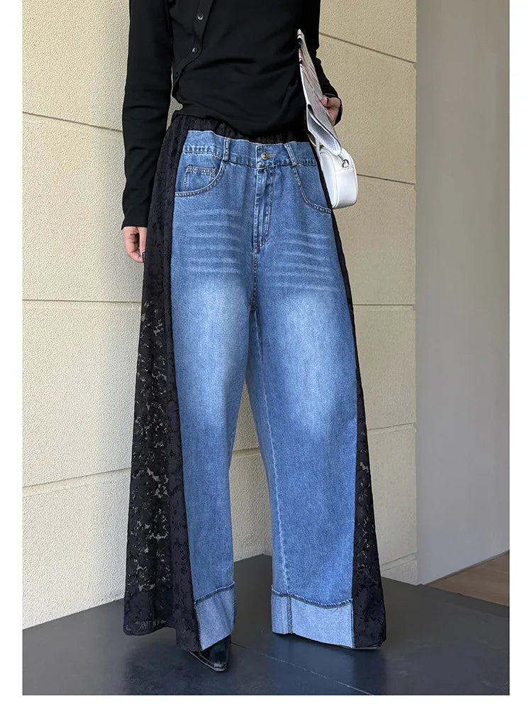 A person wearing wide-leg blue denim jeans with long black lace side panels, a black long-sleeve top, and carrying a white shoulder bag, standing against a beige paneled wall.