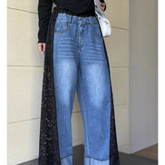 A person wearing wide-leg blue denim jeans with long black lace side panels, a black long-sleeve top, and carrying a white shoulder bag, standing against a beige paneled wall.