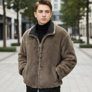 A man wearing a brown fuzzy fleece jacket with a black inner collar and black ribbed cuffs, standing indoors in front of a brown leather sofa. 