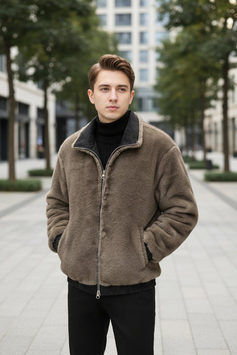 A man wearing a brown fuzzy fleece jacket with a black inner collar and black ribbed cuffs, standing indoors in front of a brown leather sofa. 