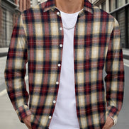 A man wearing a long-sleeve button-up plaid flannel shirt in red, beige, and black over a white t-shirt and blue jeans, standing on a city street.
