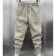 Beige baggy sweatpants hanging on a hanger against a perforated metal wall, with white sneakers below and a white border around the photo.