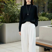 A woman wearing a black long-sleeve knit sweater with a crew neck, paired with white wide-leg pants and a silver pendant necklace. 