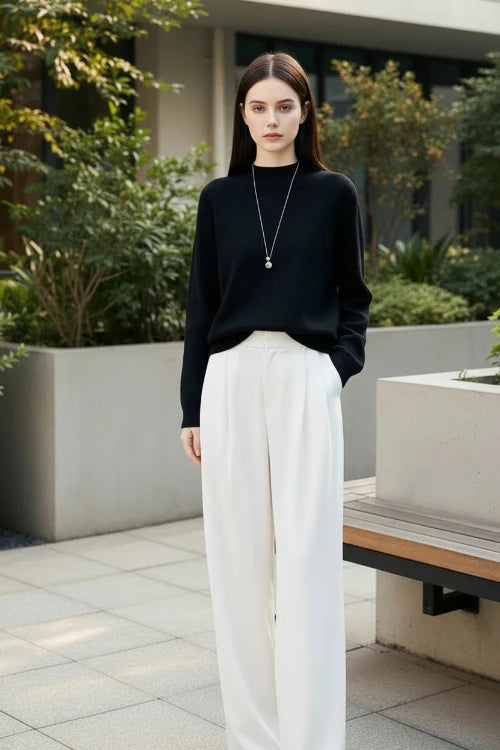 A woman wearing a black long-sleeve knit sweater with a crew neck, paired with white wide-leg pants and a silver pendant necklace. 