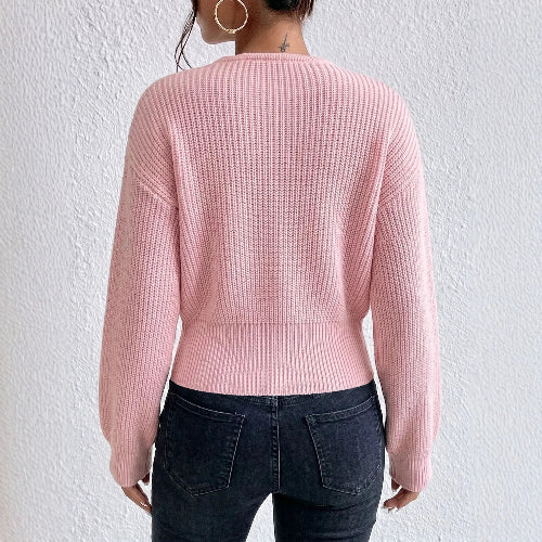 A woman shown from the back wearing a light pink ribbed knit sweater and dark blue jeans against a textured white wall.