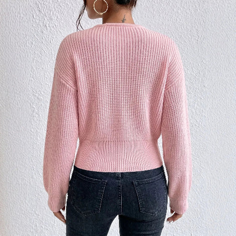 A woman shown from the back wearing a light pink ribbed knit sweater and dark blue jeans against a textured white wall.