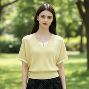 A woman wearing a pale yellow ribbed knit V-neck short-sleeve sweater over a white collared shirt, paired with a black polka-dot skirt and a black shoulder bag.