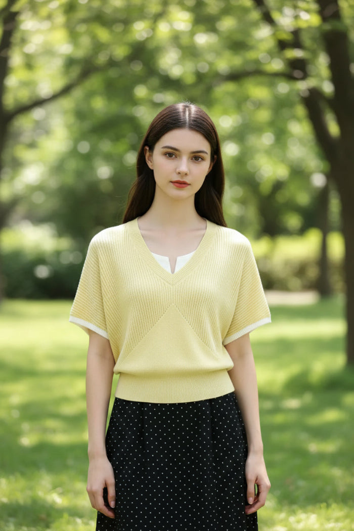 A woman wearing a pale yellow ribbed knit V-neck short-sleeve sweater over a white collared shirt, paired with a black polka-dot skirt and a black shoulder bag.