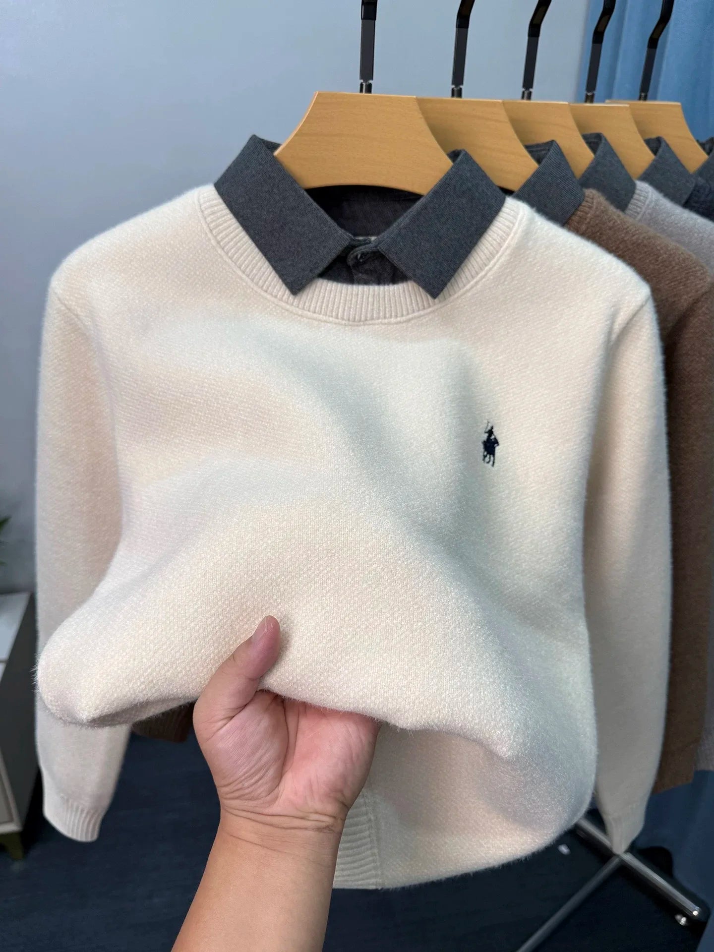 White sweater with a dark collar held by a hand, with more sweaters on hangers in the background.
