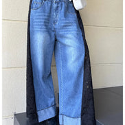 A person wearing wide-leg light blue denim jeans with long black lace side panels, styled with a black top, black pointed shoes, and a small white handbag against a beige wall.