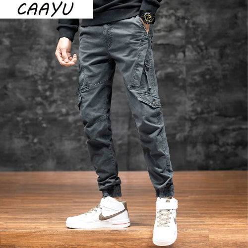Front view of a model wearing gray cargo pants with angled thigh pockets and gathered ankle cuffs, paired with white high-top sneakers against a dark wall.