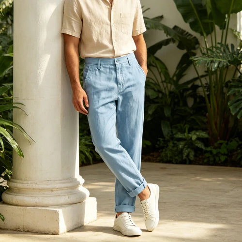 A person wearing light blue chino pants, a beige short-sleeve button-up shirt, and white sneakers, standing beside a white column in a sunlit garden setting with green plants.