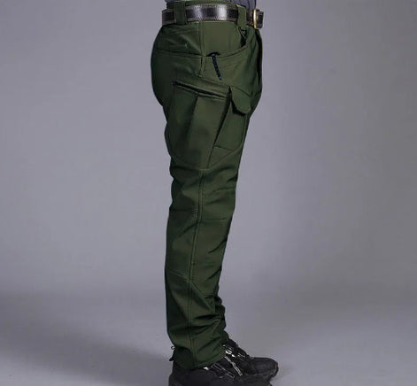 Side view of a model wearing dark olive green cargo pants with a zippered thigh pocket and flap pocket, paired with a black belt and black shoes against a gray background.