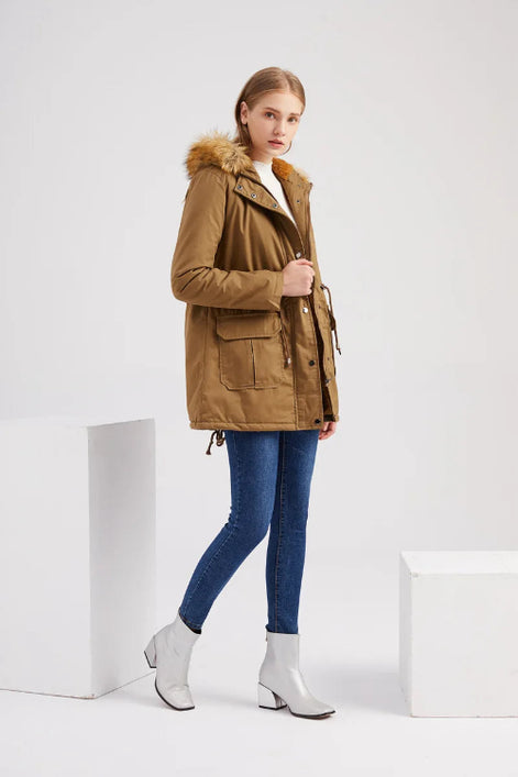 A woman models a tan hooded parka jacket with a faux-fur trimmed hood, blue skinny jeans, and white ankle boots in a minimalist studio setting with white geometric blocks.
