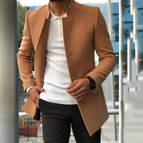 Man wearing a tan coat over a white shirt with a blurred cityscape background