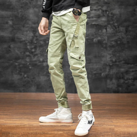 Front-angle view of a model wearing light khaki cargo pants with angled thigh pockets and gathered ankle cuffs, paired with white high-top sneakers against a dark wall.
