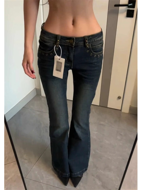 Front view of low-rise blue jeans with faded thighs, metal eyelet pocket details, and widened hems, worn with black pointed shoes.