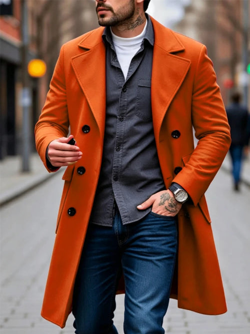 A man wearing a vibrant orange double-breasted overcoat over a dark gray button-down shirt, blue jeans, and a white t-shirt, walking on a blurred city street.