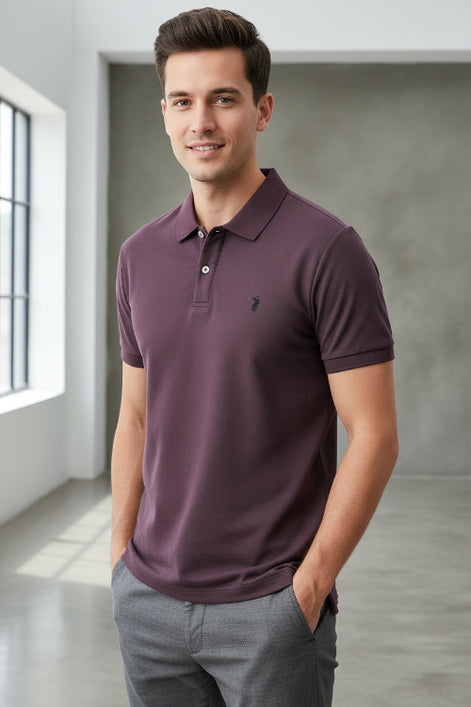 Purple polo shirt with a brand logo held by a hand, with other shirts in the background.