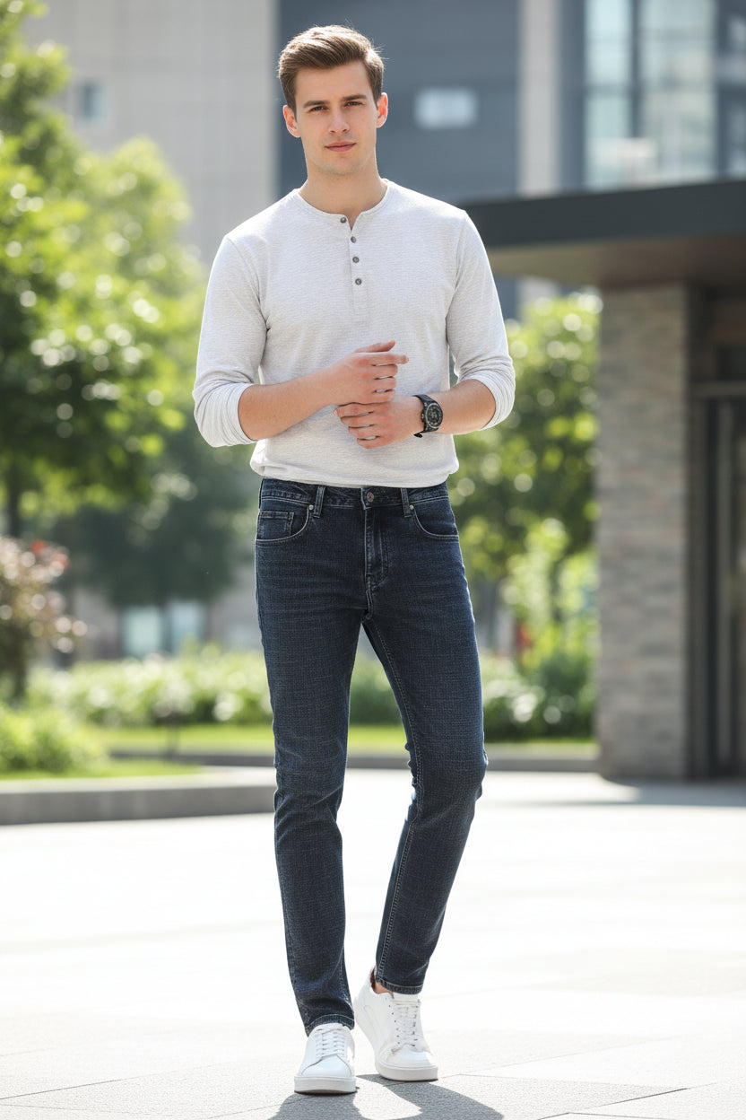 A person wearing dark gray slim-fit jeans, a brown knit sweater, and white sneakers, shown from the chest down against a plain gray studio background.