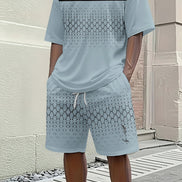 A man wearing a light blue matching t-shirt and shorts set with a geometric pattern, standing outdoors against a beige brick wall.