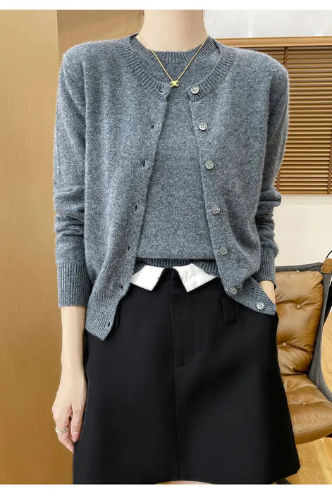 A person wearing a heather gray button-front cardigan over a white collared shirt and black shorts, standing indoors with a wooden chair and blinds in the background.