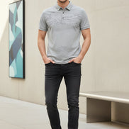 A light gray short-sleeve polo shirt with a subtle landscape print on the front and a small chest logo, shown on a black hanger with a hand holding the sleeve to demonstrate fabric texture.