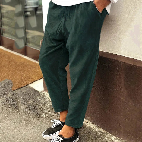 Person wearing dark green pants and black sneakers leaning against a wall.