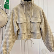 Beige and cream checked utility jacket on a hanger, showing a raised drawcord collar, chest flap pockets, front snap buttons, elasticated cuffs and a tied drawcord hem