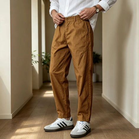 A person wearing brown relaxed-fit trousers, a white button-up shirt, and white sneakers with black stripes, standing in a hallway with beige walls and wood flooring.