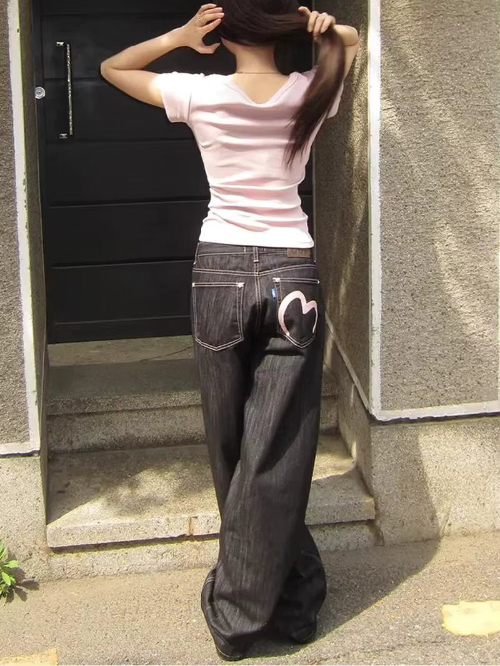 Rear view of a woman wearing dark-wash loose-fit jeans with contrast stitching and a pink heart outline on the right back pocket, standing by outdoor steps.