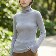 A person wearing a fitted long-sleeve turtleneck sweater in a muted gray-blue color, paired with brown bottoms, photographed indoors against a light tiled wall.