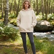 A woman modeling an oversized beige pullover hoodie with a front kangaroo pocket, photographed against a clean white studio background.