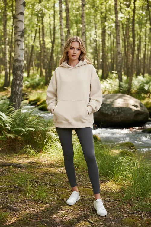 A woman modeling an oversized beige pullover hoodie with a front kangaroo pocket, photographed against a clean white studio background.