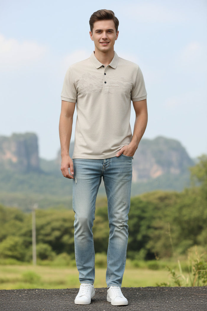 A men's short-sleeve polo shirt in a light beige or cream color, featuring a subtle, tonal landscape graphic across the chest. The shirt is displayed on a black hanger against a solid white background.