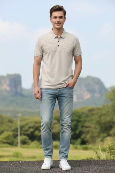 A men's short-sleeve polo shirt in a light beige or cream color, featuring a subtle, tonal landscape graphic across the chest. The shirt is displayed on a black hanger against a solid white background.