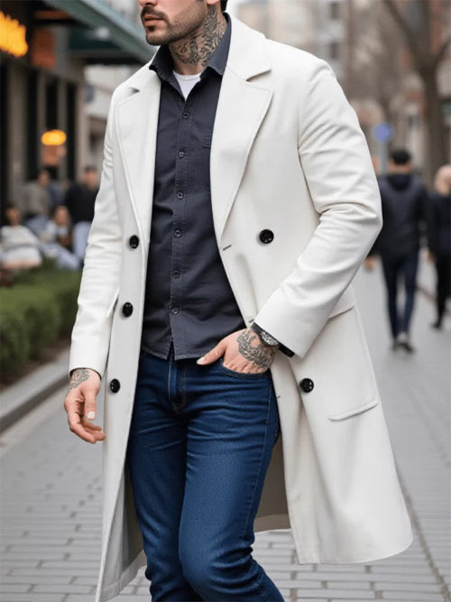 A man wearing a long white double-breasted overcoat over a dark gray button-down shirt and blue jeans, walking on a city street.