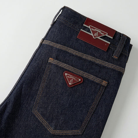 Close-up of the back waistband and pocket area of dark indigo denim jeans, showing contrast tan stitching, a red leather patch with a silver logo on the waistband, 