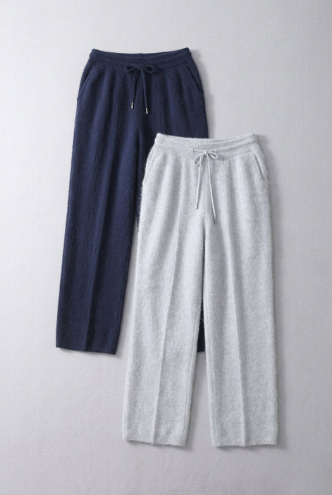 Two pairs of wide-leg knit lounge pants hanging on wooden hangers in front of a wooden shelf. The pair on the left is dark navy blue, and the pair on the right is light heather gray.