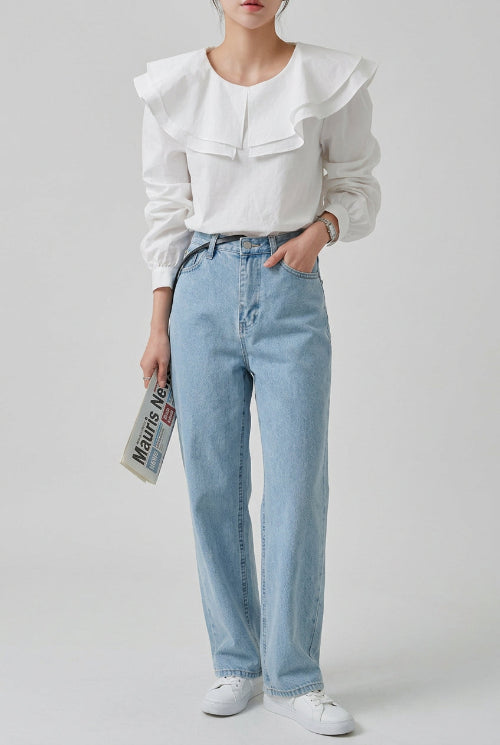 A woman wearing a white long-sleeve blouse with a dramatic oversized ruffled collar and light-wash blue jeans, 