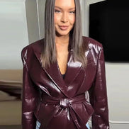 Woman wearing a glossy dark brown belted jacket with wide lapels over a white dress, holding a matching clutch on a sidewalk