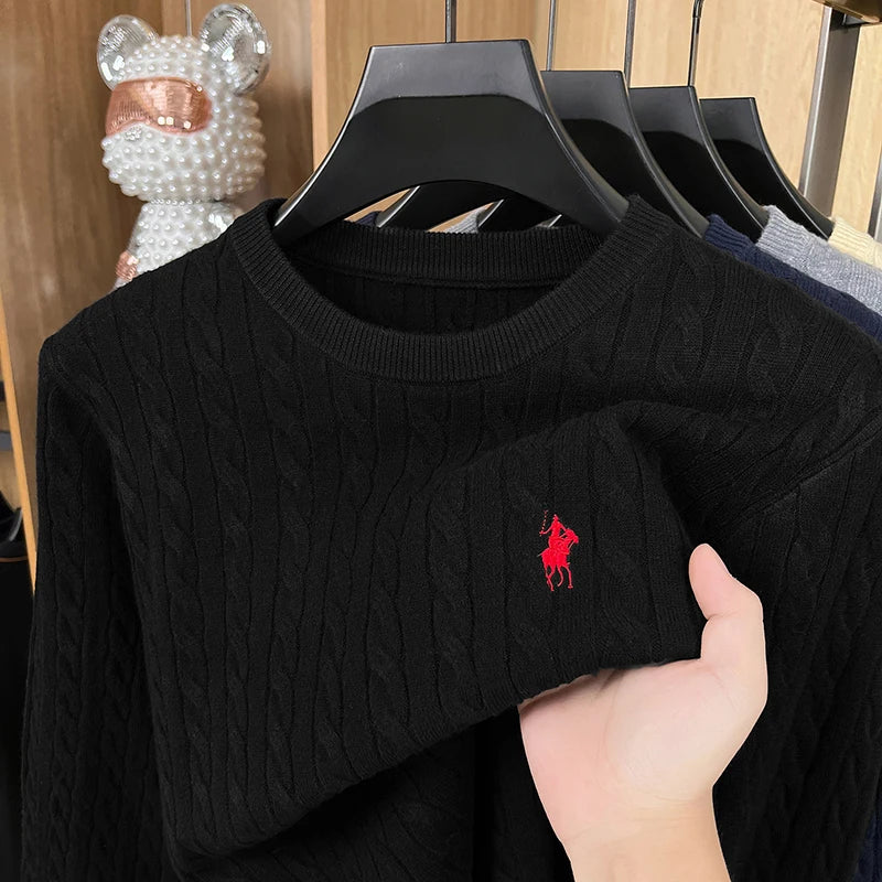 Black cable knit sweater with a red logo on a hanger, held by a hand.