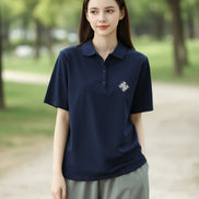 A woman wearing a navy blue short-sleeve polo shirt with a small white embroidered geometric logo on the left chest, paired with light gray pants and a brown shoulder bag.