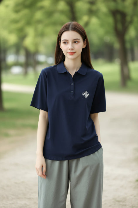 A woman wearing a navy blue short-sleeve polo shirt with a small white embroidered geometric logo on the left chest, paired with light gray pants and a brown shoulder bag.